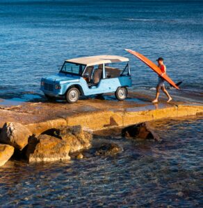 Méhari électrique eStory bleue garée sur la plage au Cap Ferret avec un homme portant une planche de surf