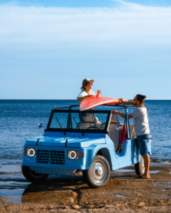 Voiture de plage électrique type Méhari avec un couple chargeant une planche de surf sur le toit, scène de plage en bord de mer, lifestyle surf et loisirs estivaux.