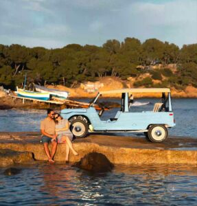 Voiture de plage électrique eStory bleue garée près de la mer en Corse avec un couple assis sur un rocher face à l’océan