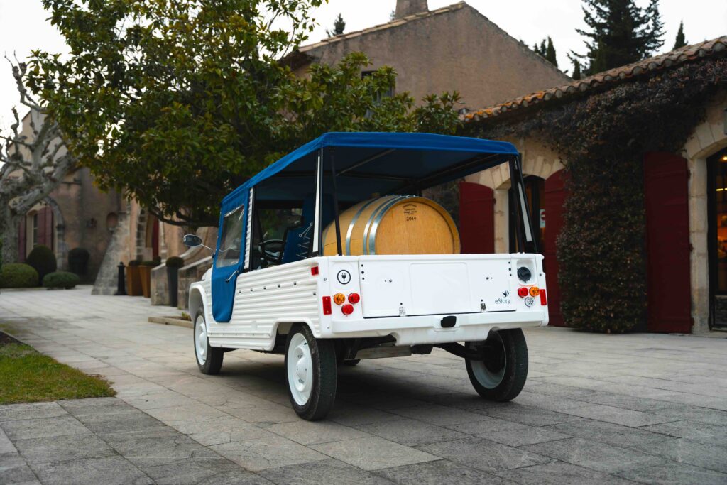 Pick-up électrique stationné dans la cour d’un domaine viticole en France, avec un grand fût ou contenant dans la benne, scène de travail dans un vignoble.